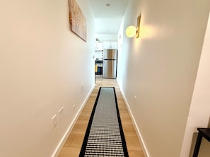 A hallway with a patterned rug leading to a kitchen area with white cabinets and stainless steel appliances. - The Golden European Flat (Charlotte)
