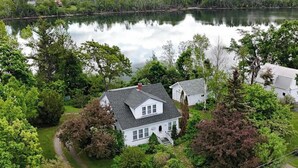 A white house with a grey roof surrounded by lush greenery and a calm lake.
