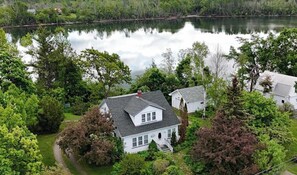 A white house with a grey roof surrounded by lush greenery and a calm lake.