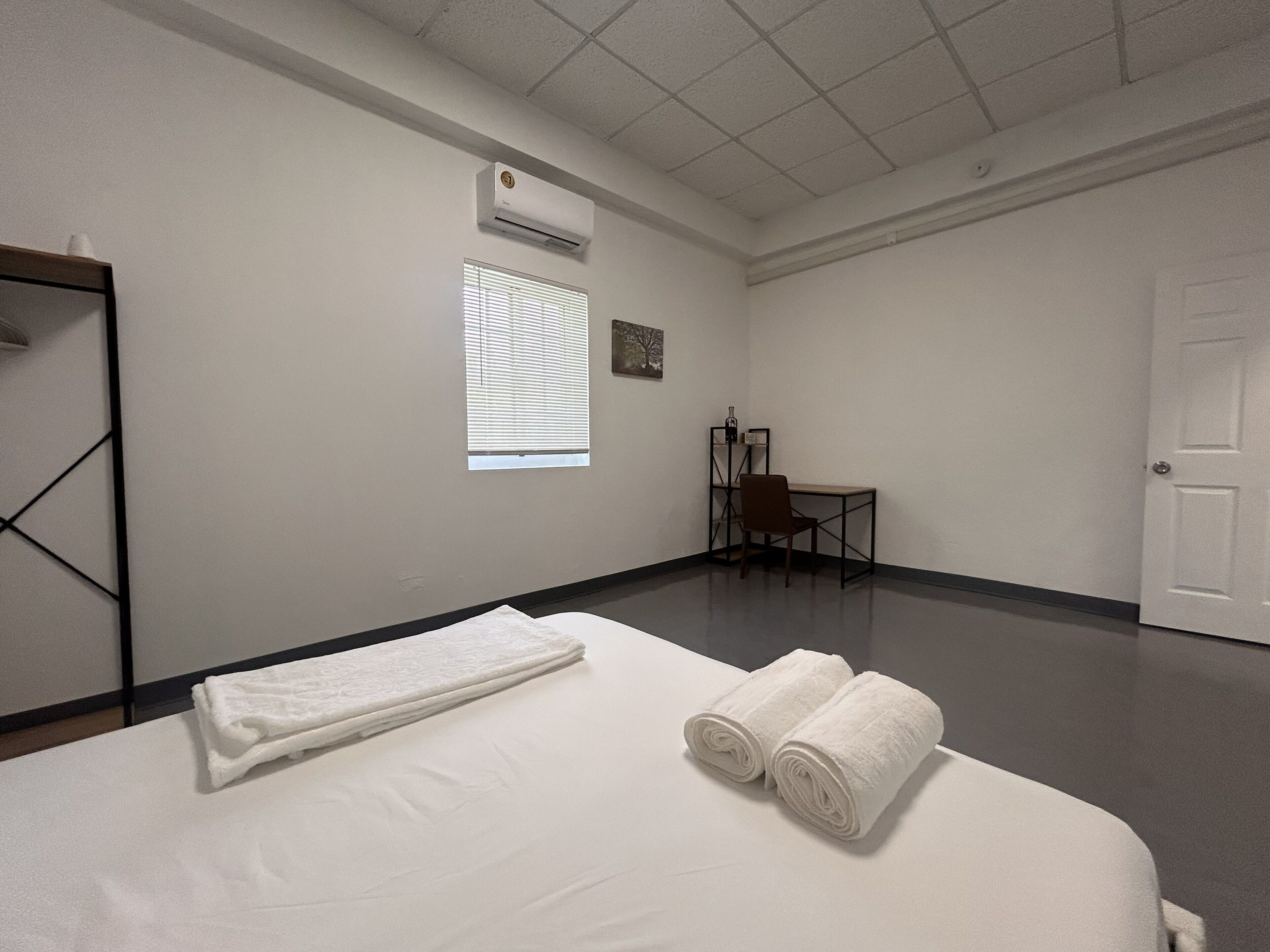 Habitación de hotel con cama, una silla, un escritorio, ventana con persianas y aire acondicionado.