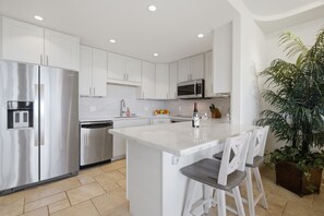 Una cocina moderna con armarios blancos, electrodomésticos de acero inoxidable, una isla central con taburetes y una planta en maceta.