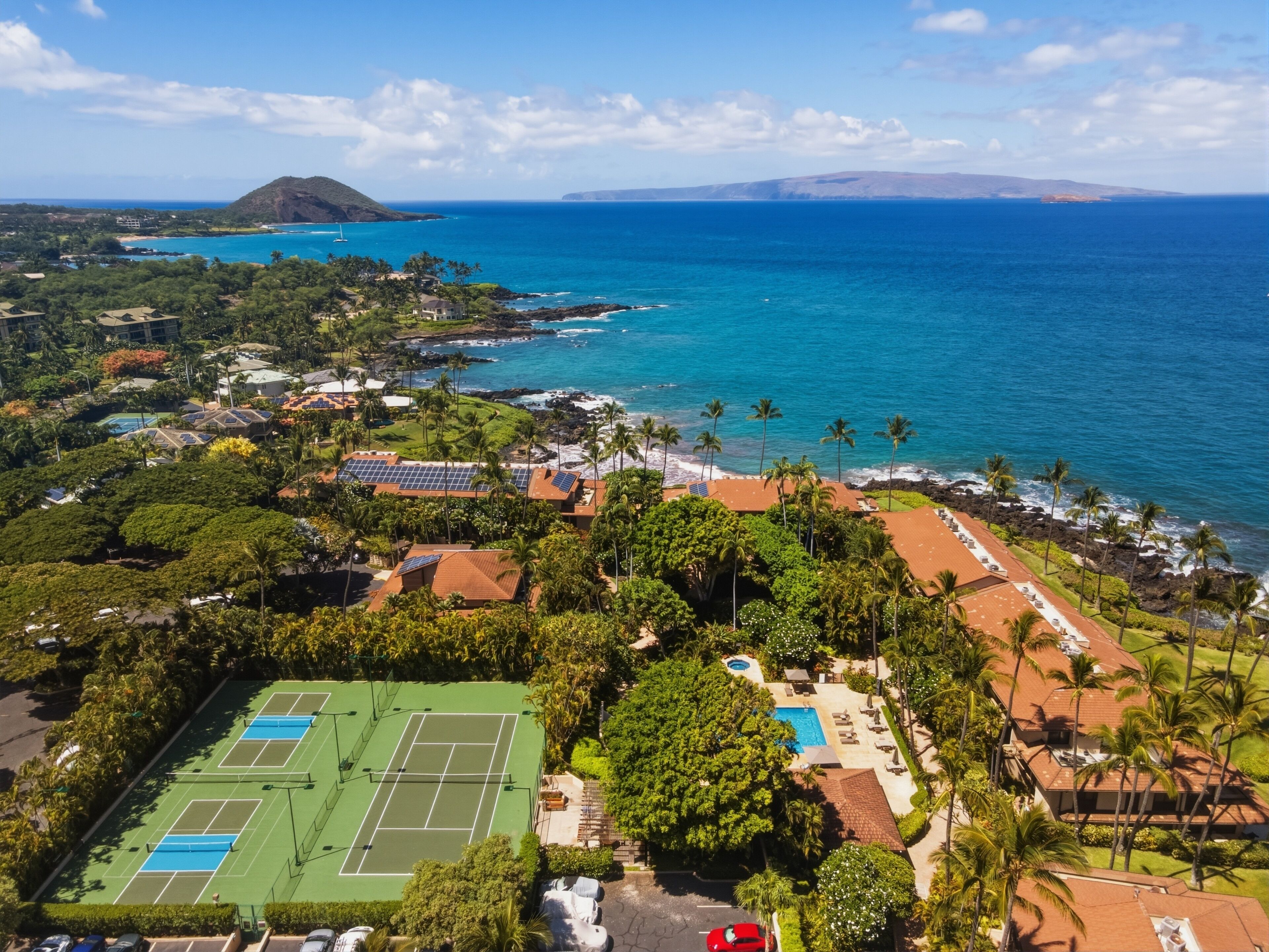 An aerial view of a coastal resort with a tennis court, pool, and ocean views.