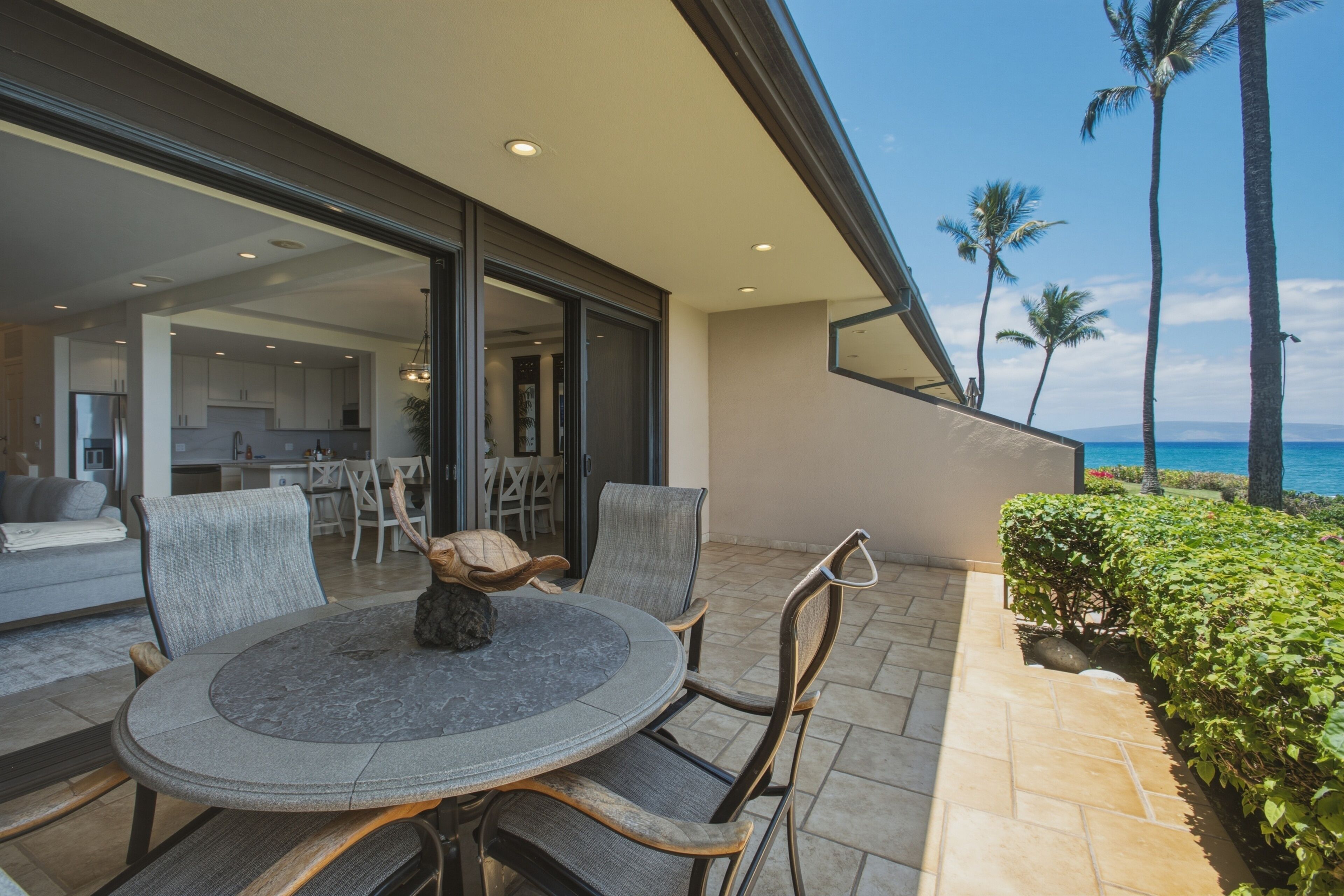 An outdoor dining area with a round table and chairs, overlooking a coastal view with palm trees and the ocean.
