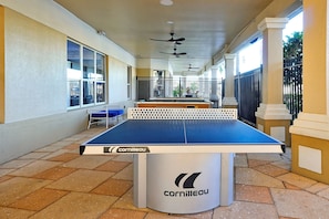 Ein Pingpongtisch mit weißem Gestell und blauer Spielfläche, befindlich in einem überdachten Außenbereich mit Fliesenboden und Säulen.