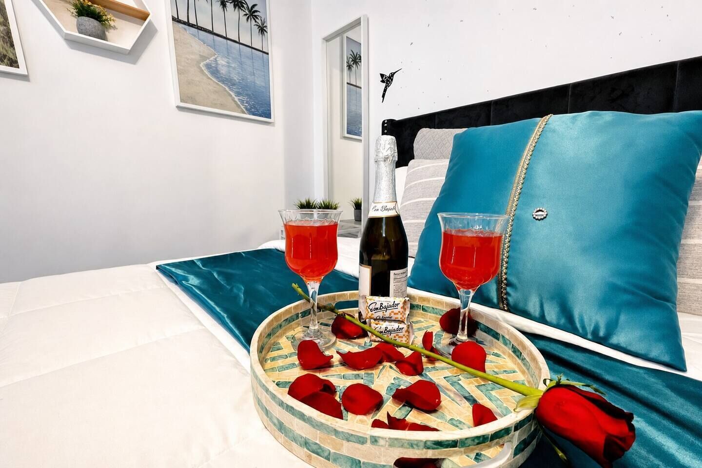 A bed with a blue pillow, a tray with rose petals, two glasses of red drink, a bottle of wine, and a framed picture on the wall.