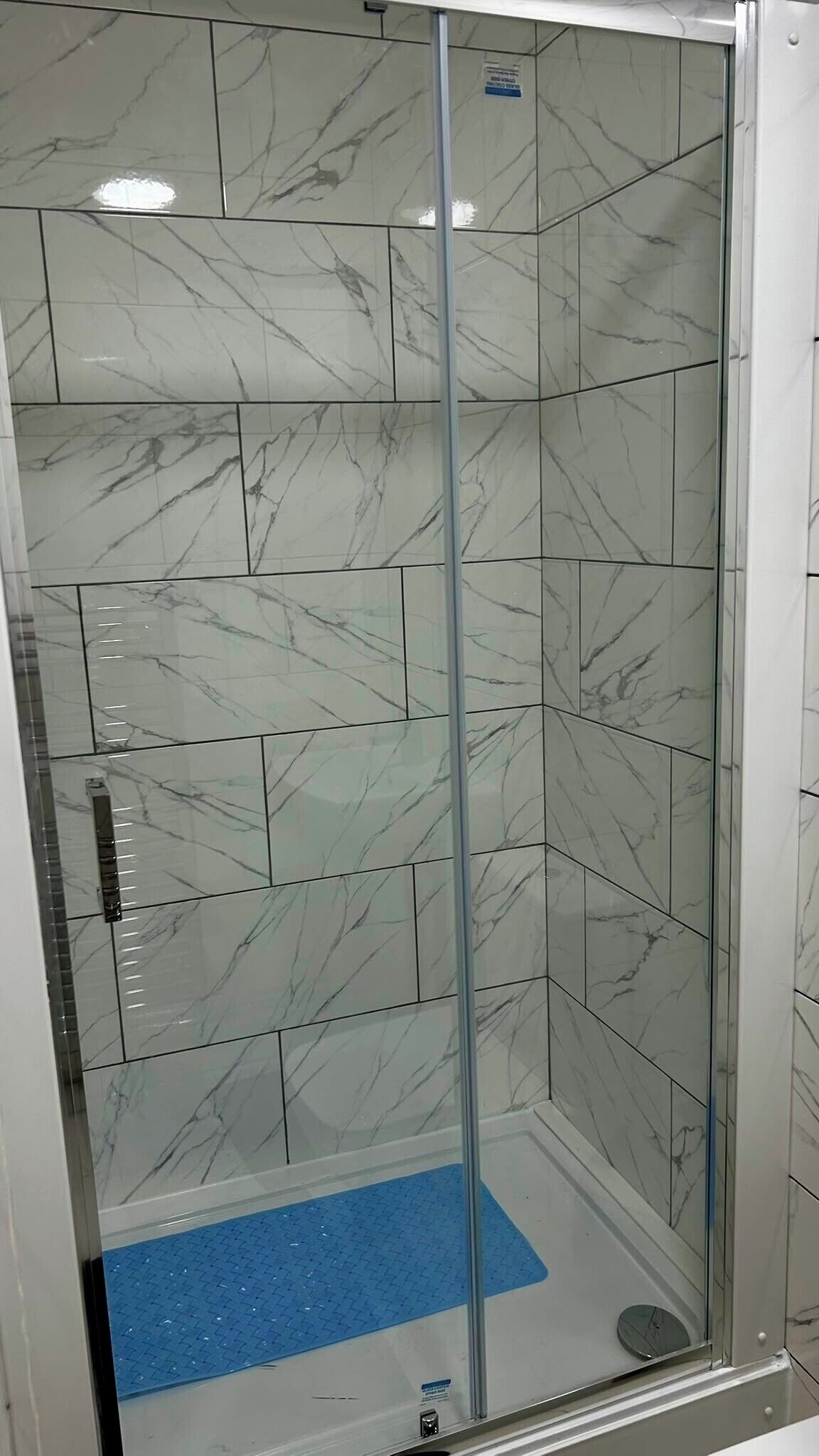 Eine gläserne Duschkabine mit Fliesen im Marmor-Design und einer blauen, rutschfesten Matte.