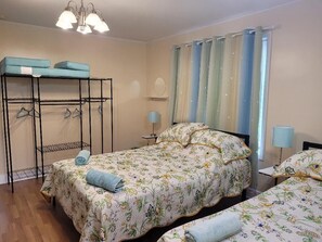 A bedroom with a bunk bed, a single bed, a bedside table with a lamp, a mirror, and a window with curtains.