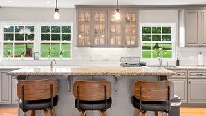 A modern kitchen with a central island, three bar stools, and a view of the outdoors through large windows.