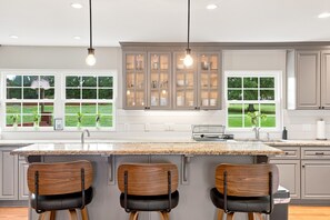 A modern kitchen with a central island, three bar stools, and a view of the outdoors through large windows.