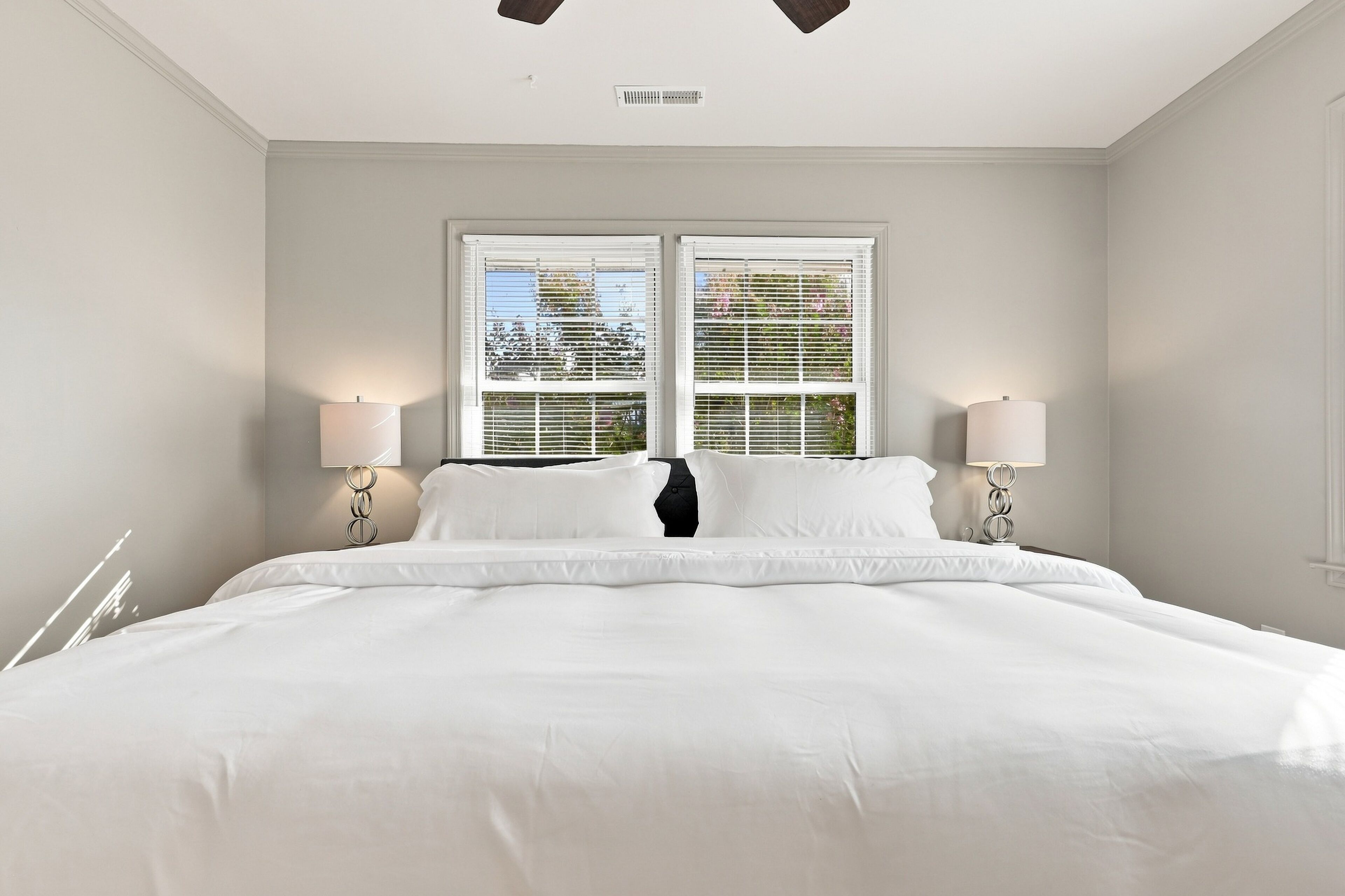 A large bed with white linens, flanked by two bedside lamps, in a room with a window and a vent on the wall.