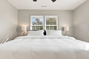 Una cama grande con ropa de cama blanca, flanqueada por dos lámparas de noche, en una habitación con ventana y conducto de ventilación en la pared.