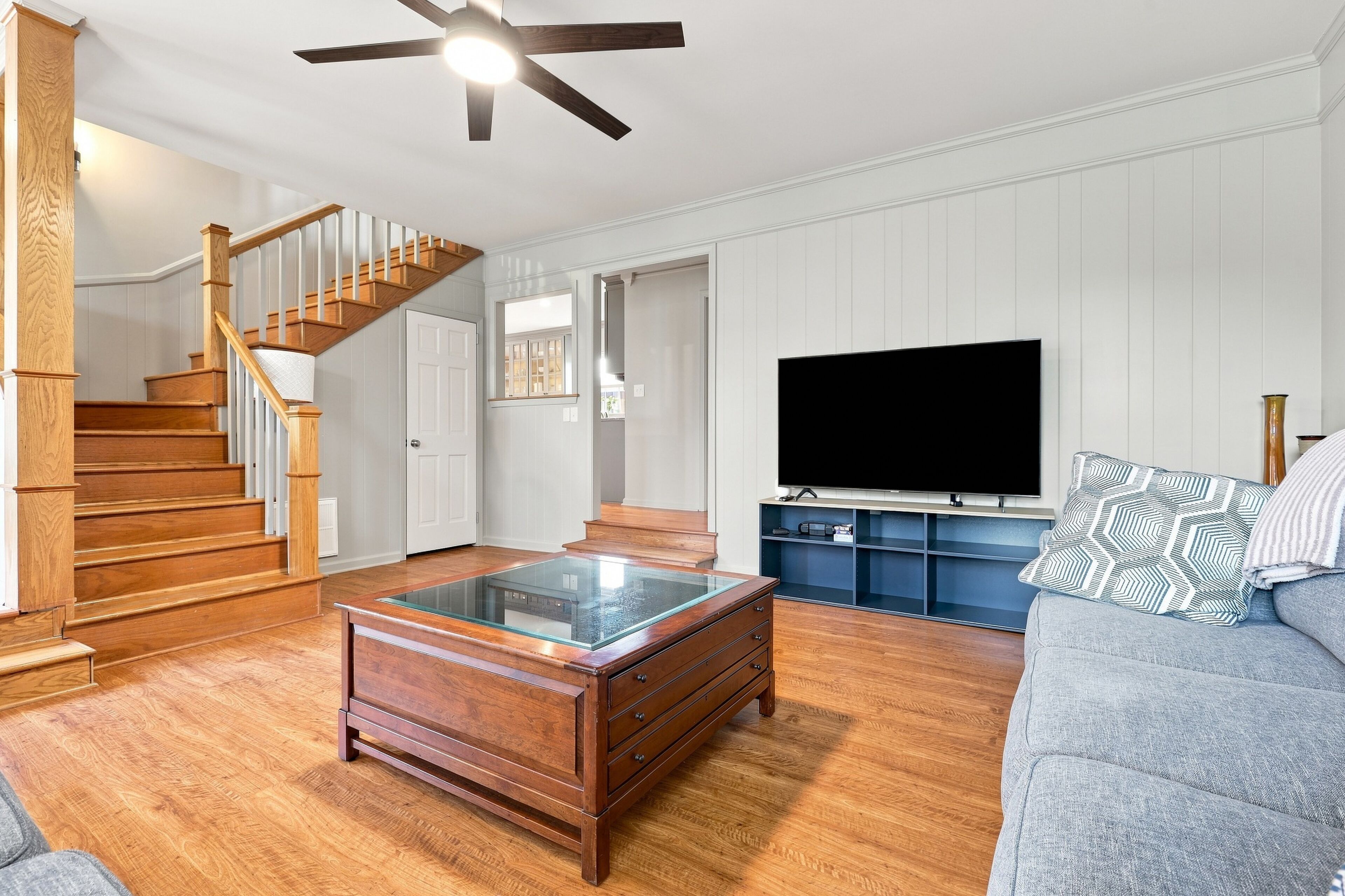 A living room with a wooden staircase, a glass coffee table, a flat-screen TV, and a grey sofa.
