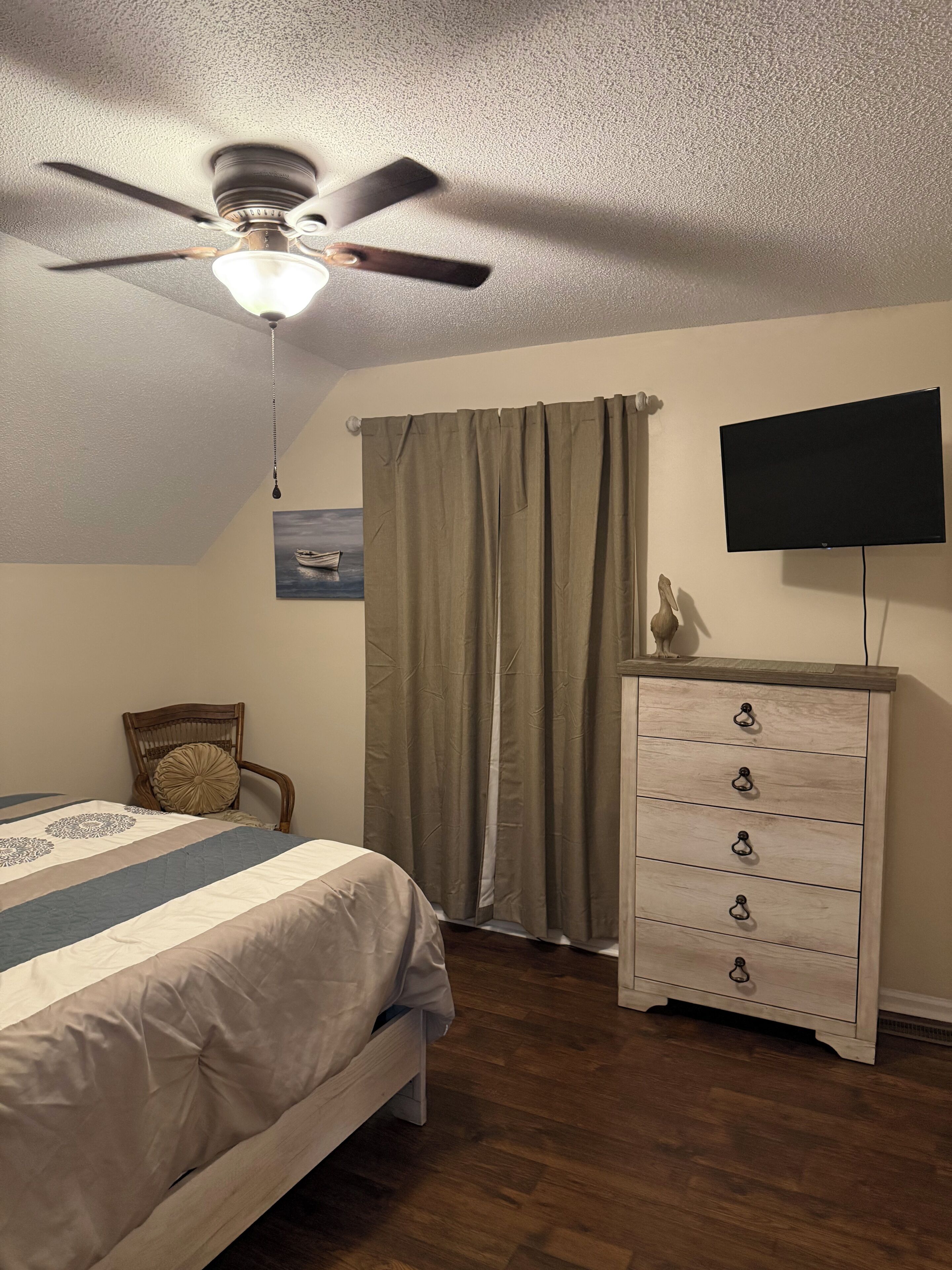 Un dormitorio con cama, ventilador de techo, cómoda, televisor y ventana con cortinas.