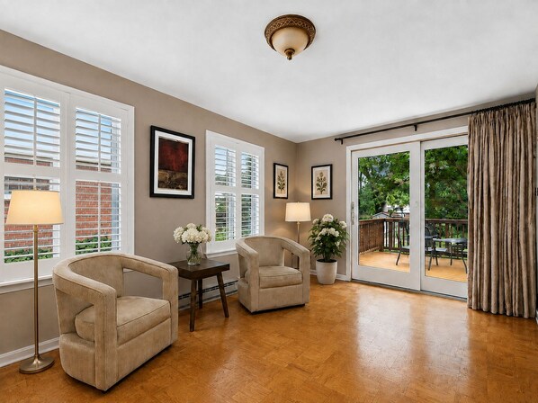 A spacious room with a large sliding glass door leading to an outdoor deck, two armchairs, a side table, and a floor lamp.