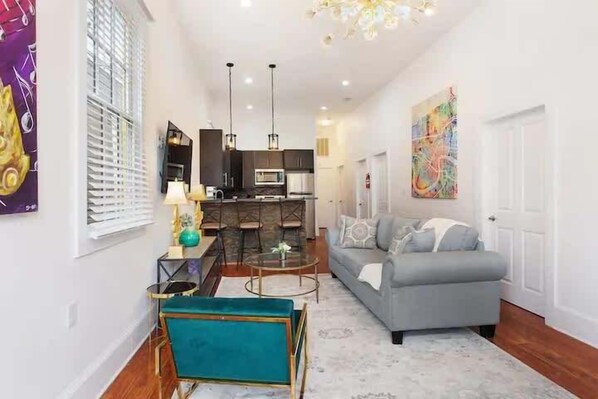 A modern living room with a sofa, a coffee table, and a TV. There is a kitchen area with a microwave and a dining table. - Spacious 4-Bed NOLA Home, Pet-Friendly (New Orleans)
