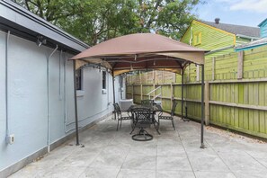 Ein Patio mit Tisch und Stühlen unter einem Vordach, ein grüner Zaun und ein Haus mit hellblauem Anstrich.