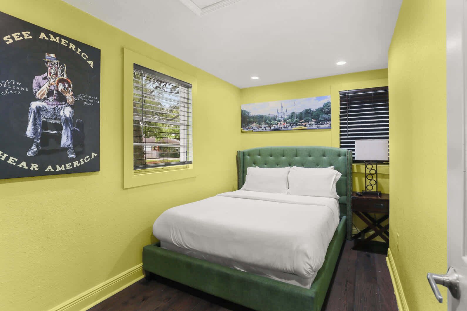 A bedroom with a green upholstered bed, a poster of a jazz musician, and a window with blinds.
