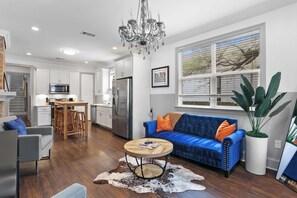 Eine moderne Küche mit Edelstahlgeräten, einem Essbereich mit Holztisch, einem blauen Sofa mit orangefarbenen Kissen und einer Kronleuchter.