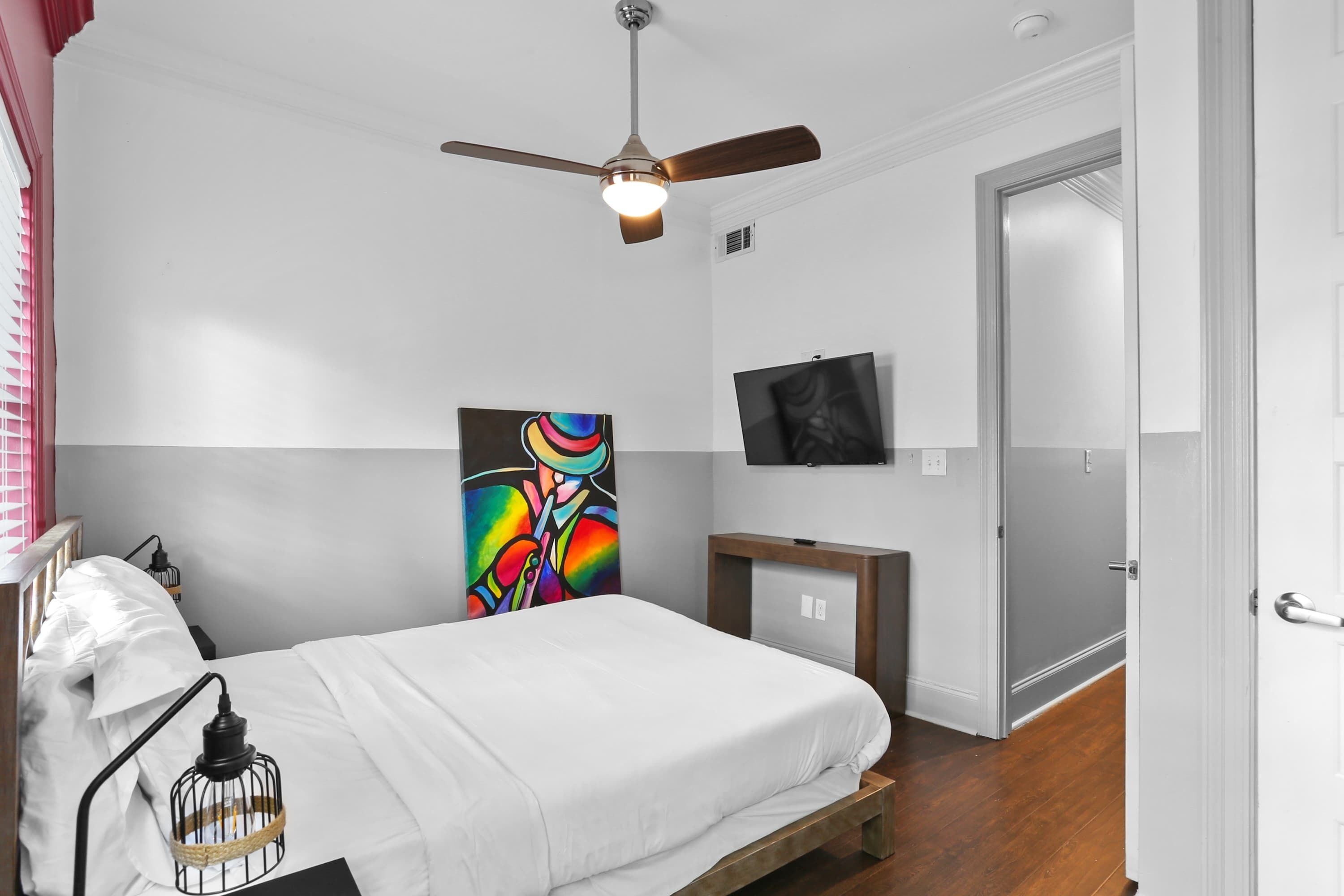 A bedroom with a bed, a ceiling fan, a TV, and a painting on the wall.