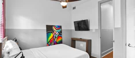 A bedroom with a bed, a ceiling fan, a TV, and a painting on the wall.
