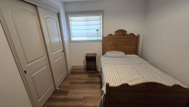 A bedroom with a wooden bed, a small bedside table, a window with blinds, and a door.
