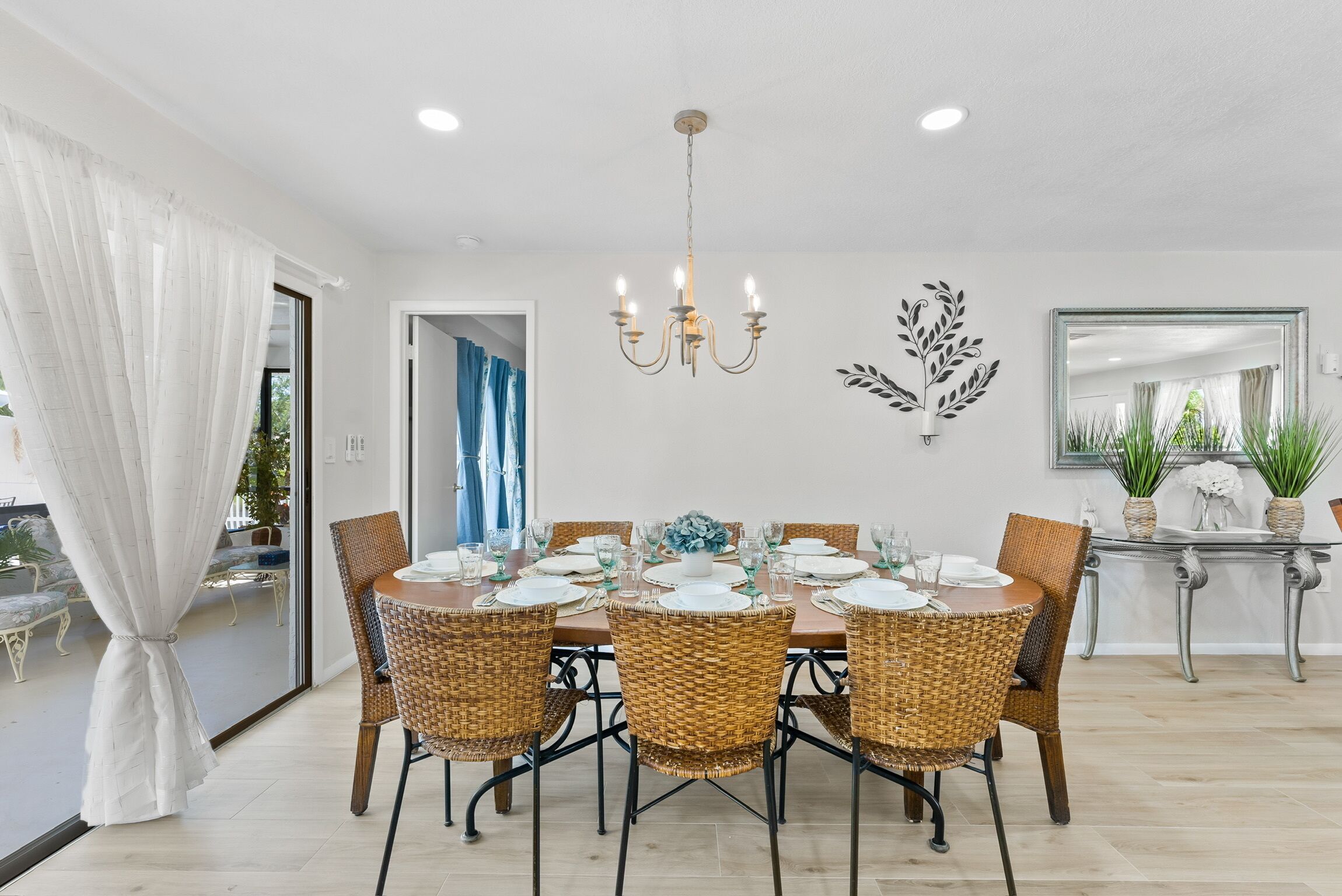 A dining area with a round table set for six, surrounded by wicker chairs, a chandelier, and a decorative wall piece.