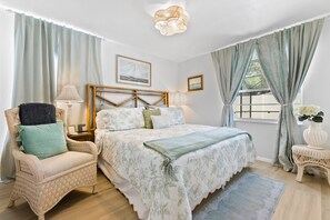 Une chambre à coucher comprenant un lit, une chaise, une table de chevet, une lampe, une fenêtre avec des rideaux et un luminaire fixé au mur.