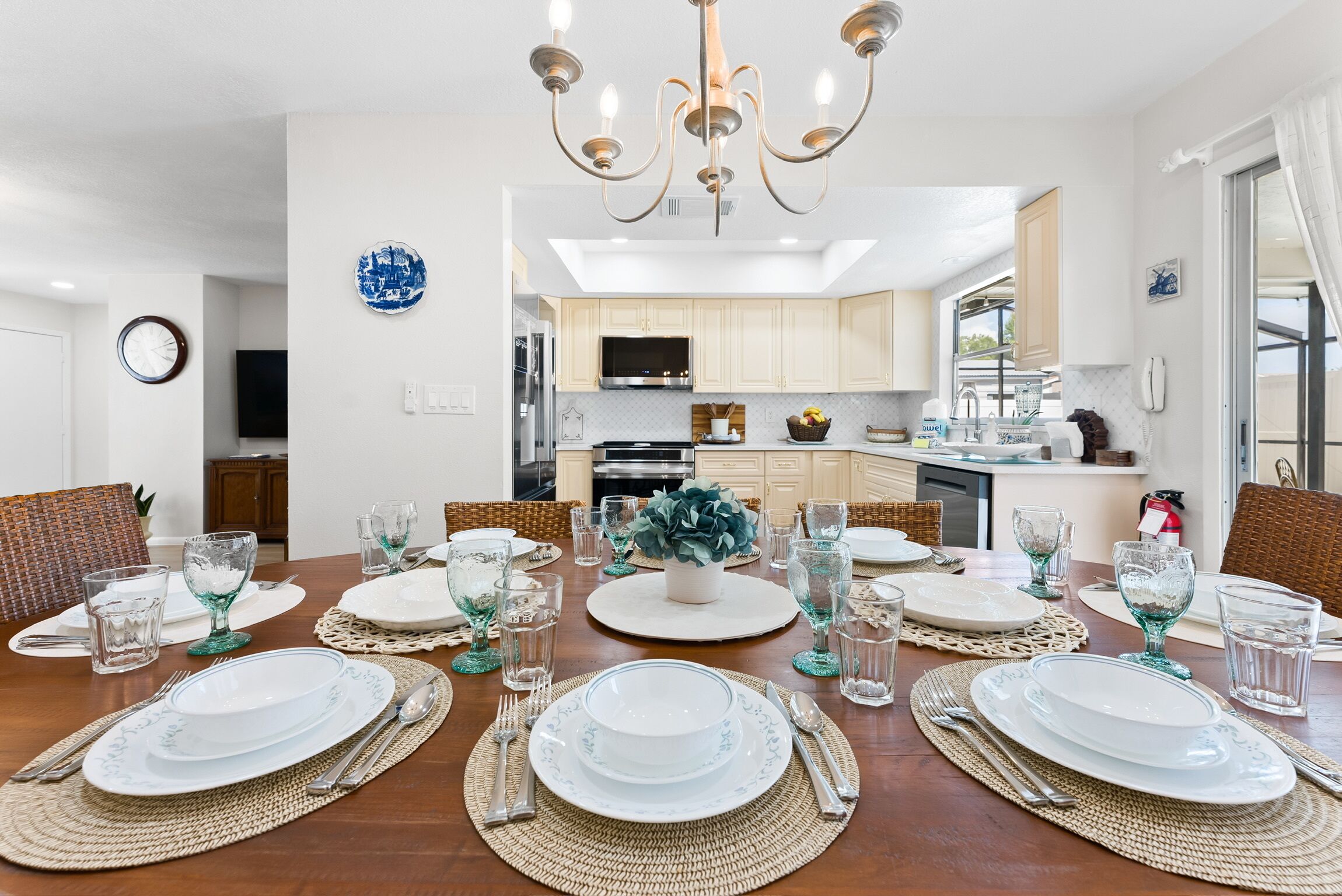 A dining area with a wooden table set for a meal, a kitchen in the background, and a chandelier above.
