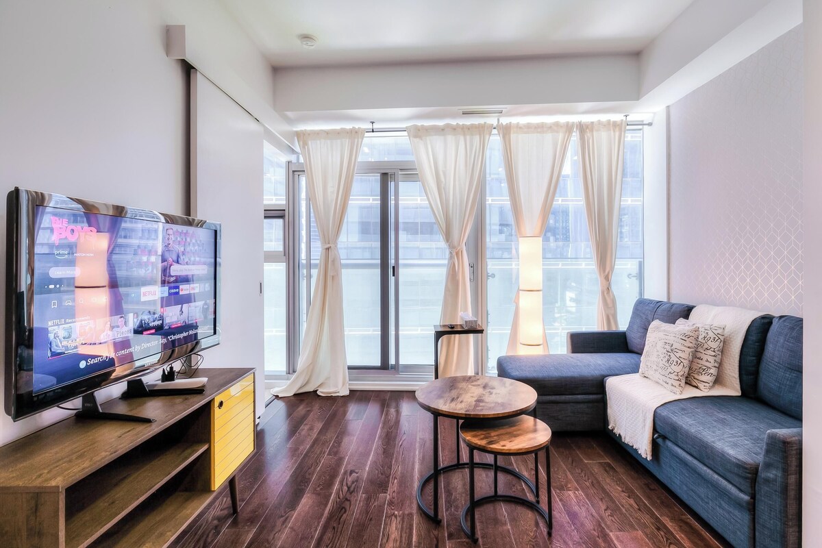 Una sala de estar moderna con una televisión de pantalla plana, un sofá, una mesita redonda y un ventanal con cortinas.