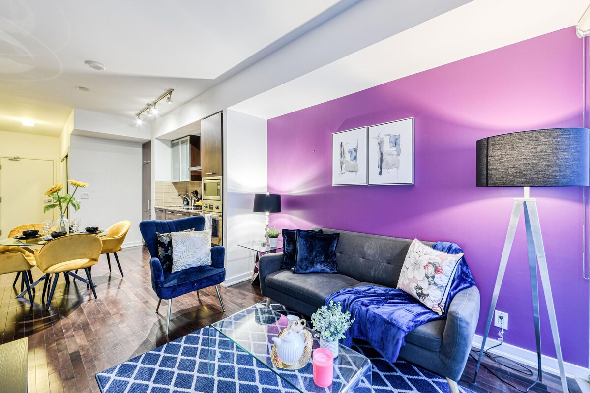 Una sala moderna con una pared de acento morada, un comedor con sillas amarillas y una cocina al fondo.