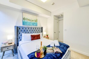 A neatly made bed with a tufted headboard, a bedside table with a lamp, a bottle of champagne, and a tray with a glass and a candle.