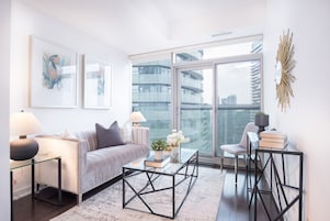 A modern living room with a sofa, coffee table, and chairs, large windows with a city view, and artwork on the walls.