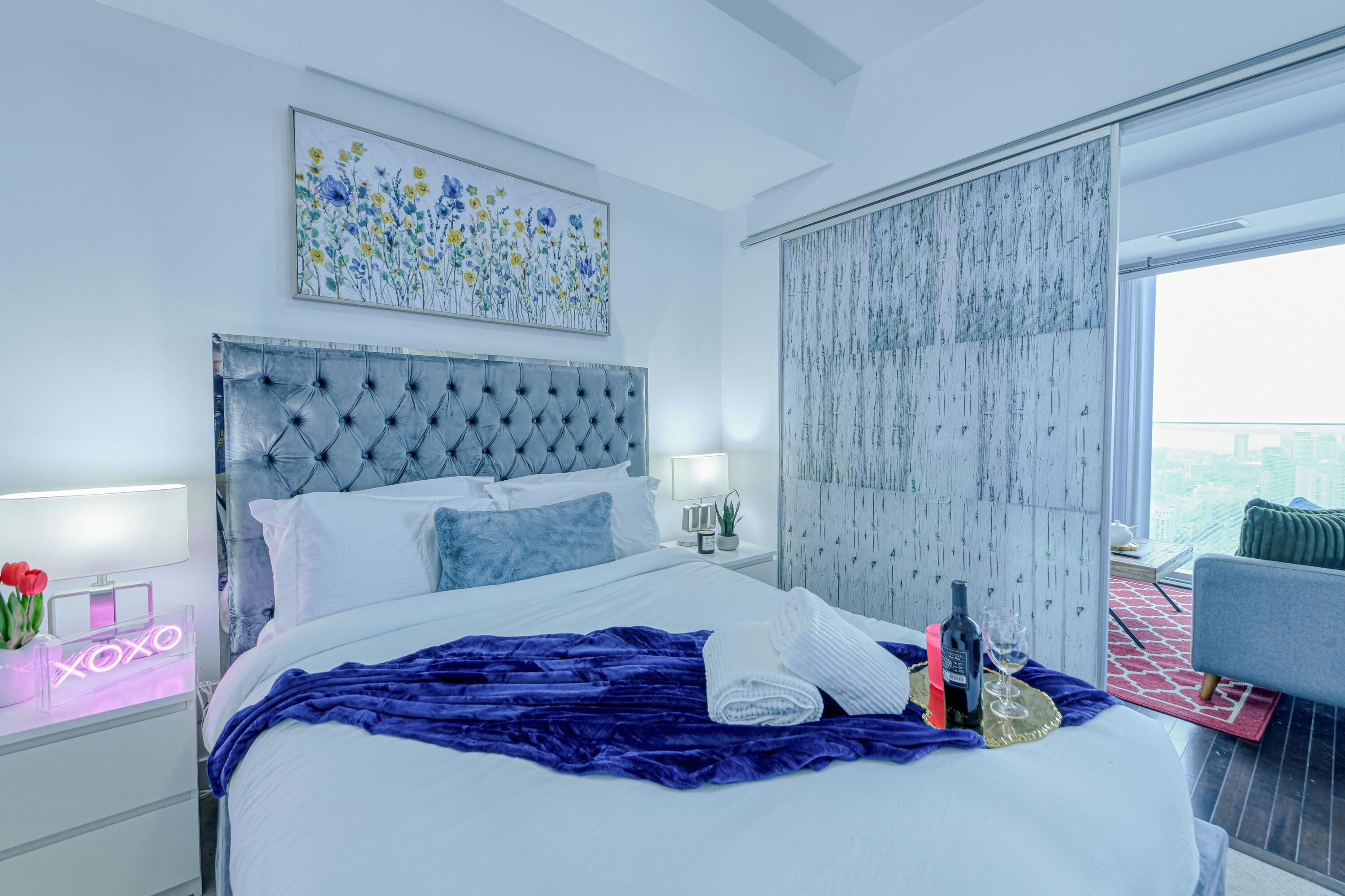 A modern hotel room with a large bed, a city view, a neon sign, and a balcony with a red and white rug.