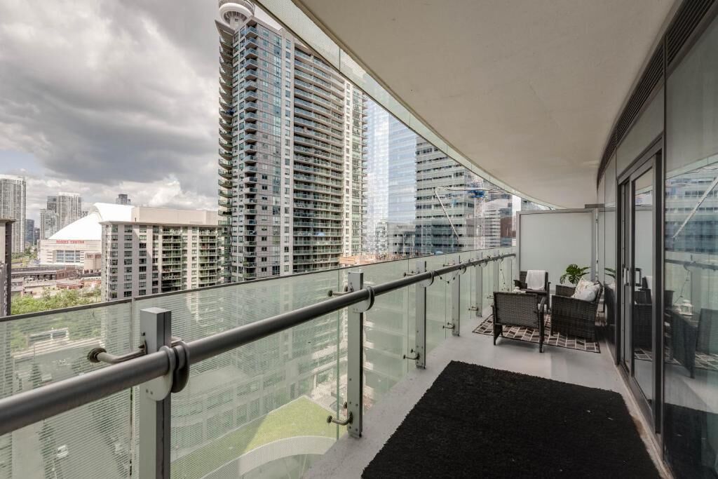 A balcony with a glass railing, a small seating area with a plant, and a view of tall buildings.