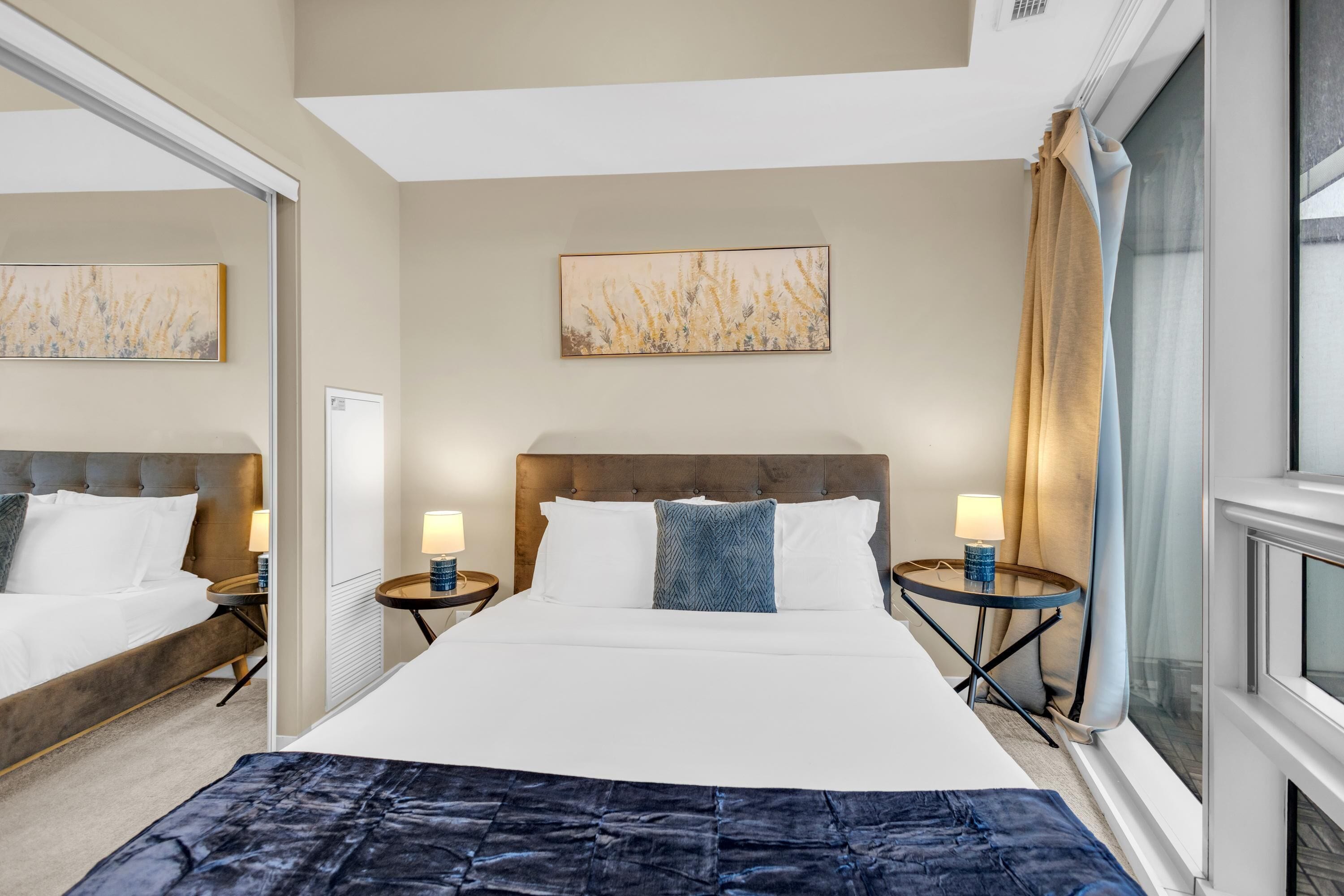 Habitación de hotel con una cama grande, dos mesitas de noche con lámparas, un cabecero, una ventana con cortinas y un cuadro en la pared.