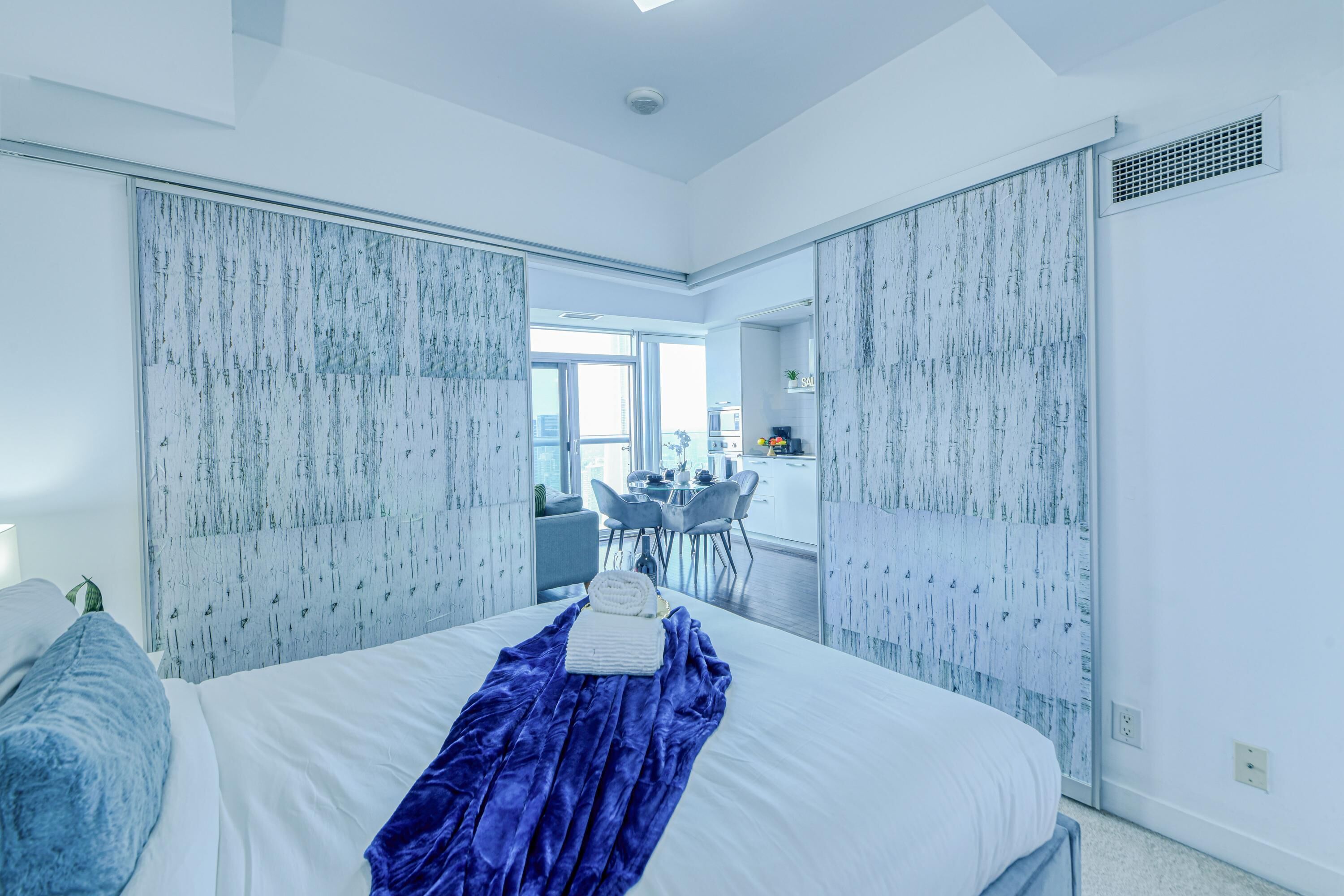 Ein modernes Schlafzimmer mit einem großen Bett, einer blauen Decke und Blick auf einen Essbereich.
