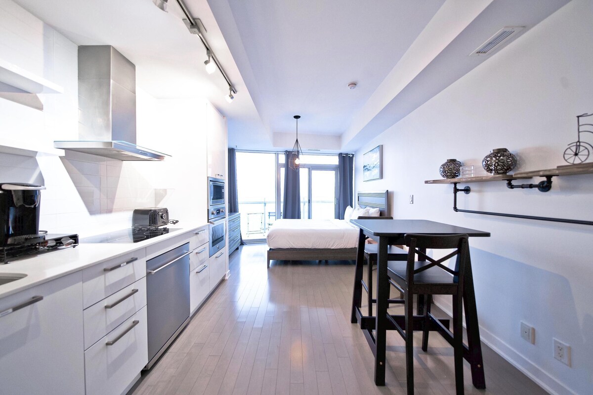 Una cocina moderna con estufa, horno y lavavajillas, un comedor con mesa y sillas, y un ventanal con cortinas.