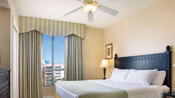 A hotel room with a bed, a nightstand, a lamp, a ceiling fan, and a window with curtains.