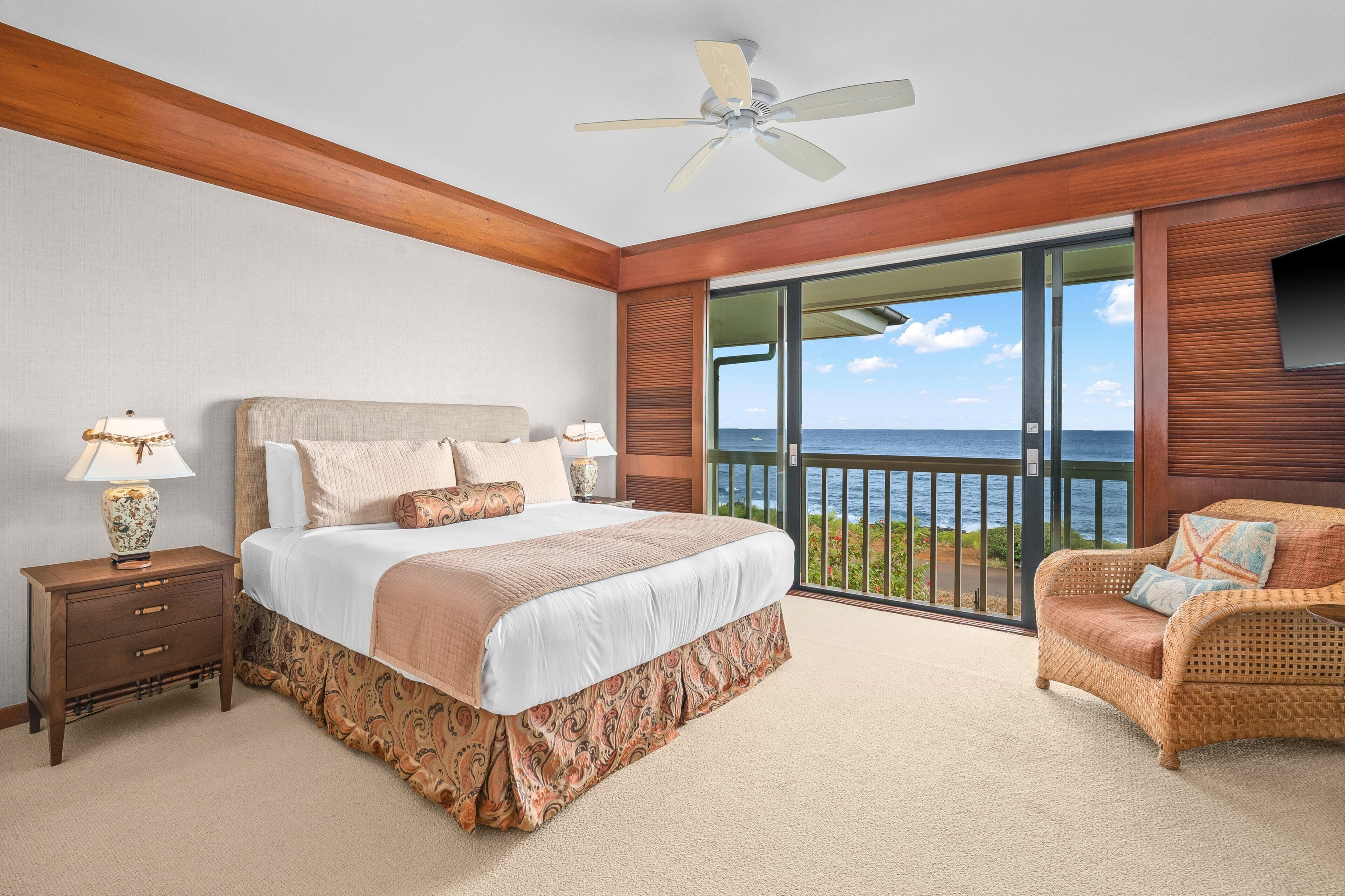 A bedroom with a large bed, a nightstand, a chair, a ceiling fan, and a view of the ocean through a sliding glass door.