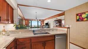 A kitchen with wooden cabinets, a stainless steel dishwasher, and a large window overlooking a scenic view. - Poipu Kapili Condo 4 by Alekona Kauai - 2BR Oceanfront (Koloa)