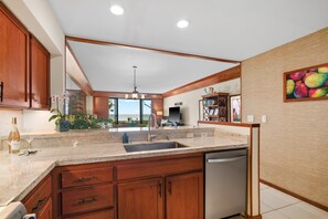 A kitchen with wooden cabinets, a stainless steel dishwasher, and a large window overlooking a scenic view. - Poipu Kapili Condo 4 by Alekona Kauai - 2BR Oceanfront (Koloa)