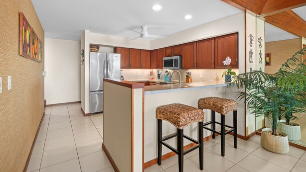 A modern kitchen with wooden cabinets, a stainless steel refrigerator, a microwave, and a bar area with stools. - Poipu Kapili Condo 4 by Alekona Kauai - 2BR Oceanfront (Koloa)