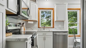 A modern kitchen with white cabinetry, a built-in oven, a dishwasher, and a window with a view of greenery. - Cedar Grove Oasis:  Huge Private Yard + Gazebo (Cedar Rapids)