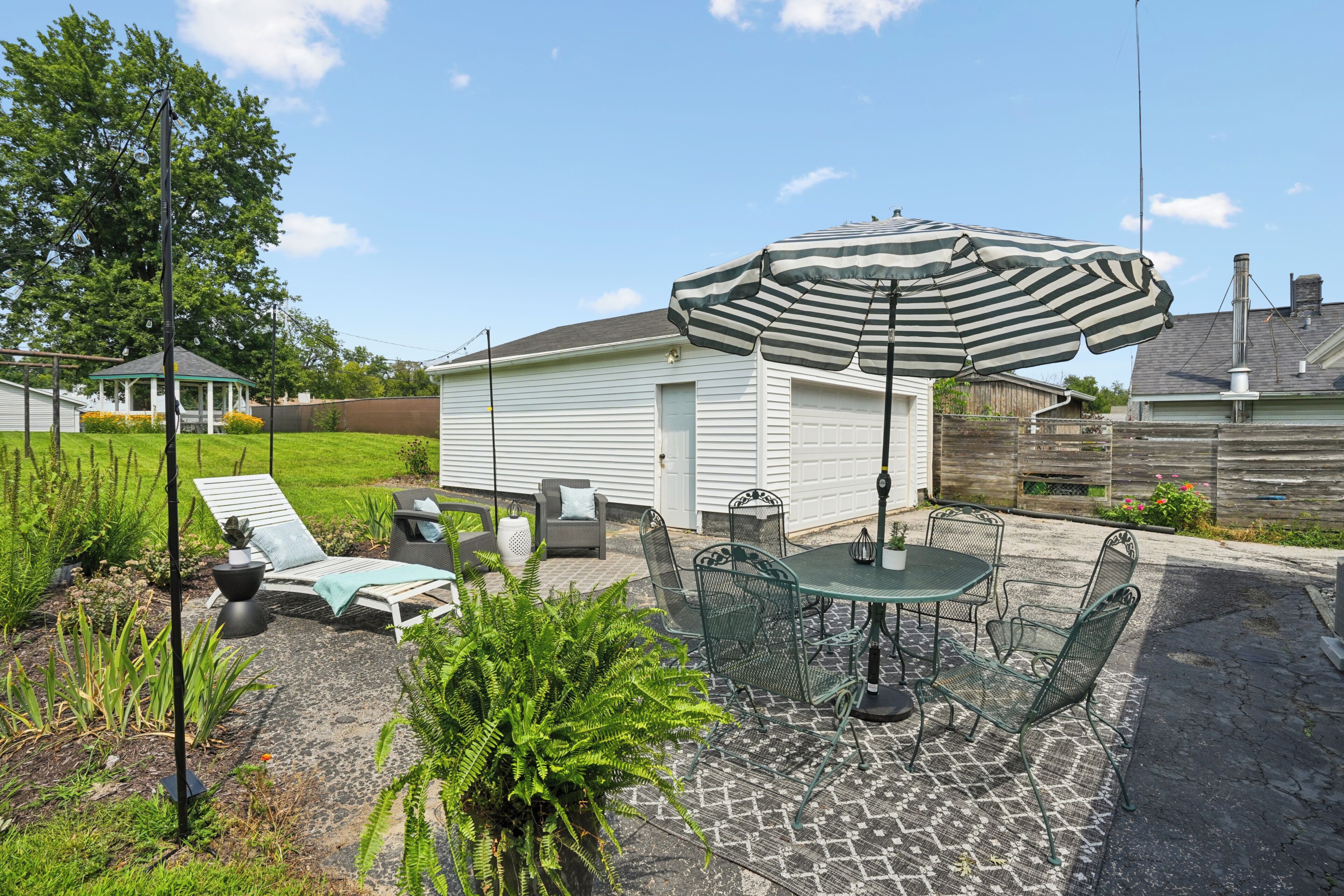 A patio with a table and chairs, surrounded by greenery and a white building.