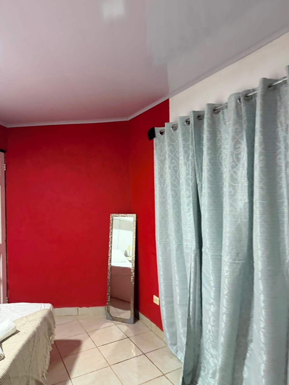 Une pièce aux murs rouges, un lit recouvert d’une courtepointe blanche, un miroir et un rideau bleu clair.