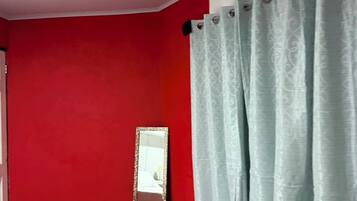 Une pièce aux murs rouges, un lit recouvert d’une courtepointe blanche, un miroir et un rideau bleu clair.
