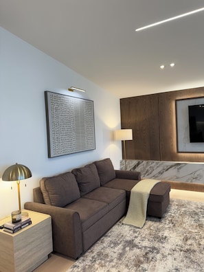 Ett modernt vardagsrum med en brun soffa, ett sidobord med böcker, en lampa, ett inramat abstrakt konstverk och ett träskåp med en tv.