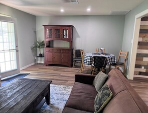 Ein Wohnzimmer mit braunem Sofa, einem Holztisch, einem Esstisch mit Stühlen und einer Holzkommode.