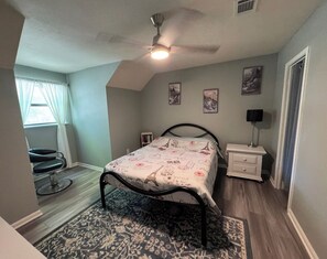 Een slaapkamer met een bed, een plafondventilator, een nachtkastje, een stoel en een raam met gordijnen.
