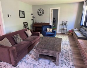 Ein Wohnzimmer mit braunem Sofa, blauem Sessel, Couchtisch aus Holz, einem gemusterten Teppich und einer Wanduhr.